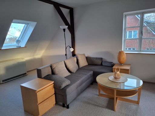 Heller Wohnraum mit grauem Sofa, Couchtisch und zwei Holzmöbeln am Fenster.