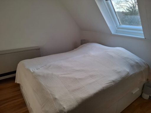 Helles Schlafzimmer mit einem Bett und schrägen Wänden unter einem Fenster.